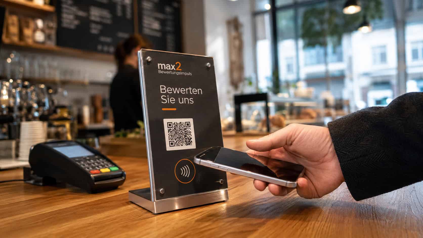 max2 BewertungsImpuls Cafe Action-Shot Gast tippt Smartphone an NFC-Aufsteller mit Google-Bewertungs-Aufforderung im Cafe